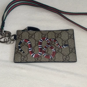 Gucci plånbok beige - Snygg plånbok från Gucci med klassiskt GG-mönster och en cool röd, vit och svart orm på framsidan. Baksidan har plats för kort och dragkedja upptill. Grön och röd rem medföljer. Gjord i skinn och canvas, perfekt för att hålla koll på kort och cash.