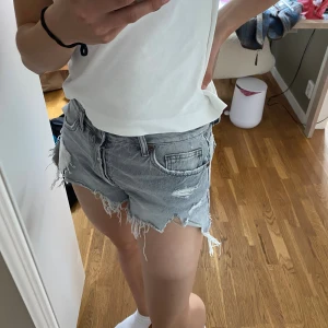 Grå slitna jeansshorts från Zara - Säljer ett par grå jeansshorts från Zara med hög midja och råa, fransiga kanter. Shortsen har flera slitningar och hål för en riktigt edgy look. Klassisk femficksmodell och knappgylf. Perfekta för sommaren och festivaler!