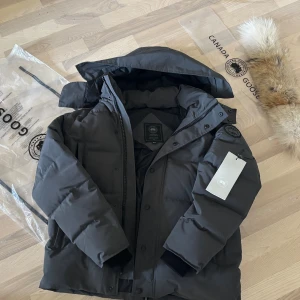 Men’s Wyndham Parka Graphite grey M/M - Säljer en svart dunjacka från Canada Goose med klassisk patch på ärmen och avtagbar huva. Jackan är rejält vadderad, har dragkedja och knappar framtill samt ribbade muddar. Perfekt för kalla vinterdagar och riktigt snygg streetstil.