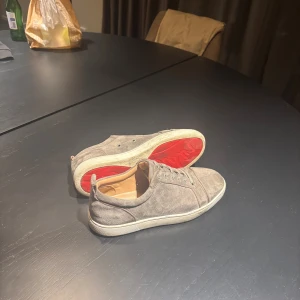 Grå mockasneakers med röd sula - Snygga grå sneakers i mocka med vit sula och cool röd undersida. Skorna har snörning och en klassisk låg profil som passar till det mesta. Insidan är beige och de har en rund tå för en avslappnad look.