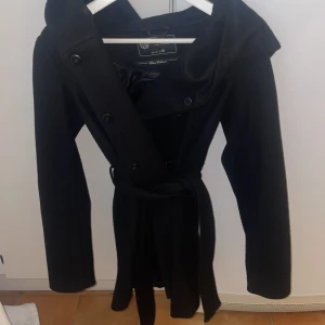 Svart kappa från Amisu med bälte - Stilren svart kappa från Amisu Classic Collection i storlek 34. Jackan har knappar framtill, ett brett bälte i midjan och en stor krage. Perfekt för dig som gillar en klassisk look med snygg passform.