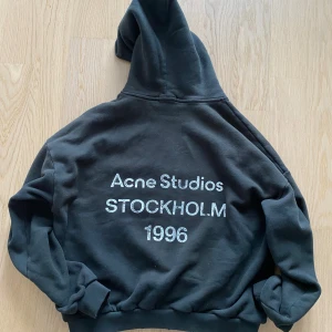Svart Acne Studios hoodie - Svart hoodie från Acne Studios med stor vit text på ryggen: 'Acne Studios STOCKHOLM 1996'. Klassisk passform med huva och ribbade muddar. Perfekt för dig som gillar stilrena streetwear-plagg med tydlig logga.
