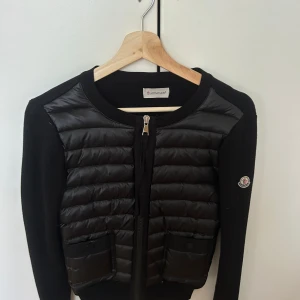Svart cardigan jacka från Moncler - Svart cardigan jacka från Moncler med quiltad front i blankt material och två fickor med knappar. Jackan har rund halsringning, dragkedja framtill och Moncler-logga på ärmen. Ärmar och rygg är i mjuk stickad kvalitet, perfekt för lager-på-lager. Jackan är äkta, (se bild 4). Köpt här på Plick för ungefär ett år sedan då den var ny men jag har knappt fått användning av den, därav säljer jag.