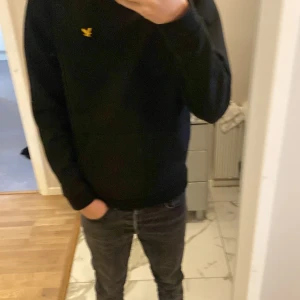 Svart hoodie från Lyle & Scott - Svart hoodie från Lyle & Scott med klassisk gul logga på bröstet. Tröjan har huva med snörning och en stor magficka. Mjuk bomullskänsla och långärmad modell, perfekt för chill dagar.