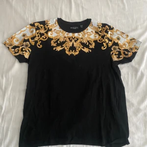 Svart t-shirt med guldmönster Brave Soul - Snygg svart t-shirt från Brave Soul med korta ärmar och ett riktigt fett barockinspirerat guldmönster över axlar och bröst. 