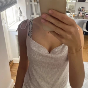 Glittrig silver topp från Bershka - Supersnygg glittrig silverfärgad topp från Bershka med tunna axelband som knyts på axlarna. Toppen har en draperad v-ringning och är croppad med resår nertill. Perfekt för att sticka ut med en cool och trendig look.