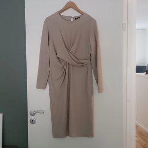 Beige långärmad klänning Stockh LM - Stilren beige klänning från Stockh LM i mjuk polyesterblandning. Långärmad modell med snygg drapering framtill och rund halsringning. Klänningen har en rak passform och slutar strax under knäet. Perfekt för dig som gillar minimalistisk och elegant stil.