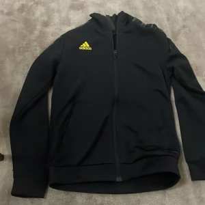 Svart Adidas jacka med dragkedja - Svart sportig jacka från Adidas med hel dragkedja framtill och gul logga på bröstet. Tillverkad av återvunnen polyester (Parley Ocean Plastic). Jackan har långa ärmar och tryck på ena ärmen. Perfekt för träning eller chill.