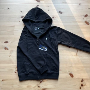 Svart hoodie från Polo Ralph Lauren - Svart hoodie från Polo Ralph Lauren med vit broderad logga på bröstet. Slim fit-modell i mjuk bomull, perfekt för en chill och stilren look.
