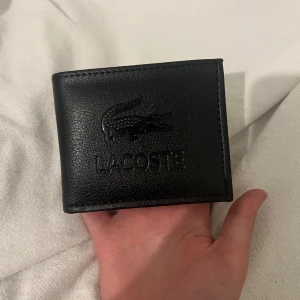 Svart plånbok från Lacoste i skinn - Snygg svart plånbok från Lacoste i skinn med präglad krokodil-logga på framsidan. Plånboken har flera kortfack, fönster för ID och plats för sedlar. Stilren och praktisk design som passar perfekt i fickan.