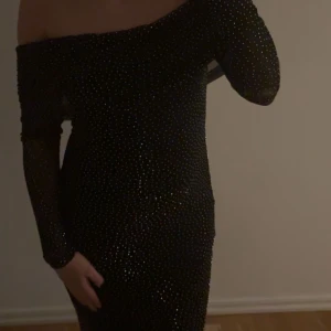 Svart glittrig nyårs klänning  - Supersnygg svart långklänning med glittriga detaljer och mesh-ärmar. Offshoulder-modell som sitter tight och markerar figuren. Perfekt för dig som vill sticka ut med en elegant och trendig look. Klänningen har lång ärm och är täckt av små glittrande stenar.