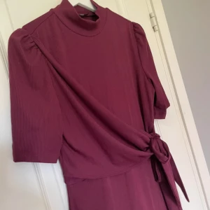 Vinröd klänning med knyt i midjan - Supersnygg vinröd klänning med halvpolo och korta puffärmar. Klänningen har ett knytband i midjan som ger en fin siluett och är tillverkad i ett mjukt, ribbat material. 