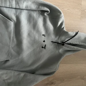 Grå Off-White hoodie med blått tryck - Säljer en grå hoodie från Off-White med svart logga framtill och stort blått och svart grafiskt tryck på ryggen. Tröjan har huva med svarta snören och klassisk känguruficka. Perfekt för dig som gillar streetwear och vill sticka ut.