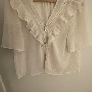 Vit blus med volang från Zara - Supersöt vit blus från Zara med volangdetaljer längs v-ringningen och pärlemorknappar framtill. Blusen har korta, luftiga ärmar och ett diskret prickigt mönster i tyget. Supersöt, och har knappt använt💞