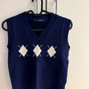 Mörkblå stickad väst från Zaful - Mörkblå stickad väst från Zaful med klassiskt rutig argylemönster i vitt framtill. Västen har v-ringning och breda ribbade kanter vid hals och ärmhål. Perfekt att styla över en skjorta för en preppy look. Västen är ny och har aldrig använt.