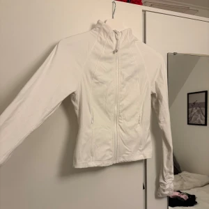 Vit träningskofta - Vit vindjacka med hög krage och hel dragkedja framtill. Jackan har två fickor med dragkedja och en figurnära passform. Långärmad och tillverkad i ett lätt, syntetiskt material som passar perfekt för en aktiv livsstil.