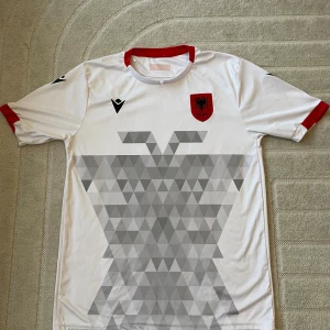 Albaniens vit fotbollströja från Macron - Säljer en officiell vit Albanien fotbollströja från Macron med röd krage och röda muddar. Framsidan har ett grått geometriskt mönster och Albaniens emblem på bröstet. Materialet är lätt och andas, perfekt för match eller träning.