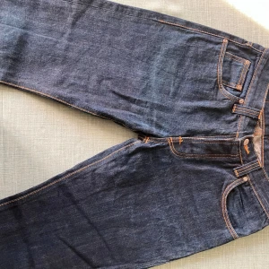 Nudie Jeans Straight Sven dry denim - Mörkblåa Nudie Jeans Straight Sven i rå denim med klassiska orange sömmar och femficksdesign. Jeansen har rak passform och är tillverkade i 100% bomull. Snygga detaljer på bakfickan och stilren look som funkar till allt.