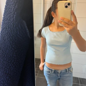 Ljusblå ribbad topp med volangkant - Supersöt ljusblå topp från Lindex med ribbad struktur och volangkant vid bordet halsringning, ärmslut och nederkant. Modellen är croppad och har korta ärmar samt en fyrkantig ringning. Perfekt för dig som gillar en enkel men ändå söt stil.