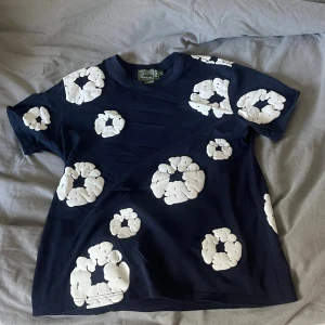 Mörkblå t-shirt från Denim Tears S - Snygg mörkblå t-shirt från Denim Tears med unika vita blommiga mönster över hela plagget. Klassisk passform med rund hals och korta ärmar. Tillverkad i mjuk bomull, perfekt för dig som vill sticka ut med en cool och modern stil.