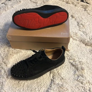 Christian Louboutin svarta sneakers med nitar - Svarta sneakers från Christian Louboutin med ikoniska röda sulor och svarta nitar på tån. Skorna har snörning och är tillverkade i mocka och skinn. Snygg och edgy design som sticker ut, perfekt för dig som gillar statement pieces.