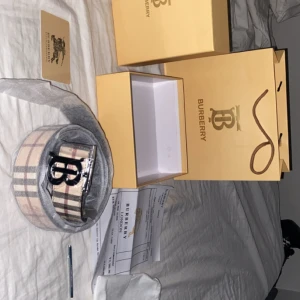Burberry rutig bälte med logga - Säljer ett snyggt Burberry-bälte med klassiskt beige, svart och rött rutmönster och en stor metallspänne med Burberrys B-logga. Perfekt accessoar för att lyfta din outfit med en lyxig touch. Kommer med originalförpackning och certifikat. Du kan också vända sidan på bältet så det blir helsvart.