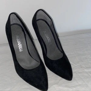 Svarta pumps från Aquarius med klack - Snygga svarta pumps från Aquarius i mocka-look med spetsig tå och hög klack. Klassisk och stilren design som passar perfekt till att lyfta vilken outfit som helst. Skorna har en elegant siluett och är tillverkade i ett mjukt material som ger en lyxig känsla.