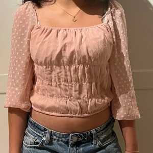 Ljusrosa blus med puffärmar - Supersöt ljusrosa blus med croppad passform och trekvartsärmar i transparent tyg med små prickar. Blusen har rynkade detaljer och fyrkantig ringning, perfekt för en trendig look.