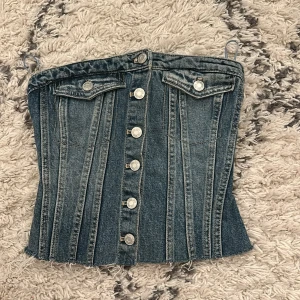 Jeanskorsett från Zara med knappar - Säljer en snygg jeanskorsett från Zara i klassisk blå denim. Toppen har silverfärgade knappar framtill, två fejkade bröstfickor och råa kanter nertill för en edgy look. Baksidan har smock för extra stretch och passform. Perfekt att styla med kjol eller byxor.