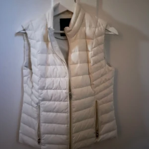 Vit dunväst från Rockandblue - Snygg vit dunväst från Rockandblue med quiltad design och dragkedja framtill. Västen har två fickor med dragkedja (alla applikationer är i guld) och hög krage. Perfekt att ha över en hoodie eller tröja för extra värme!