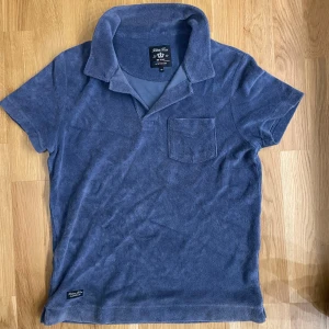 Blå frotté pikétröja Robbie Moor XS - Säljer en blå pikétröja från Robbie Moor i mjuk frotté. Tröjan har klassisk krage och en bröstficka. Perfekt för chill dagar eller när du vill ha en avslappnad men snygg look.