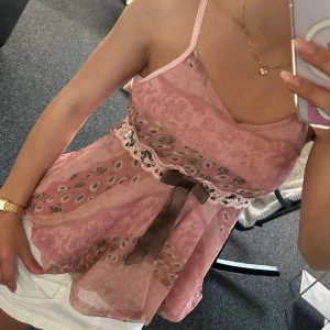 Rosa mönstrad topp med rosett - Söt rosa topp med tunna axelband och v-ringning. Toppen har ett blommigt och prickigt mönster, dekorerad med spets och paljetter i midjan samt en brun rosett framtill. Fladdrigt material som ger en luftig känsla.