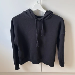 Fin svart hoodie från Pull&Bear - Svart hoodie från Pull&Bear i storlek XS. Klassisk modell med huva och snörning framtill, tillverkad i mjuk bomulls- och polyestermix. Perfekt för en chill och avslappnad stil. Långärmad och med ribbade muddar vid ärmslut och nederkant.