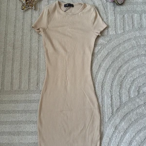 Beige ribbad kortklänning från F&F - Supersnygg beige ribbad kortklänning från F&F med rund halsringning och korta ärmar. Klänningen har en tight passform och är perfekt för dig som gillar en enkel men stilren look. Tillverkad i mjuk bomull som känns skön mot huden.