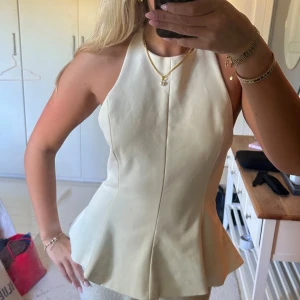 peplumtop från Zara - Supersnygg champagnefärgad peplumtop från Zara med halterneck och markerad midja. Toppen är ärmlös och har en lätt glansig finish, dragkedja i sidan och en stilren, elegant siluett. Perfekt för dig som vill sticka ut med en chic och modern look.