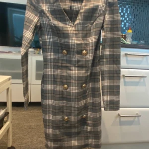 Rutig grå kortklänning från Amisu - Snygg kortklänning från Amisu i grått och vitt rutigt mönster. Klänningen har lång ärm, markerad midja och dubbla rader med guldknappar framtill. V-ringad omlottlook och figurnära passform ger en chic och modern stil.