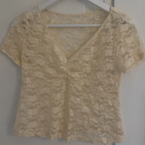 Ljus gul spetsblus med v-ringning - Söt ljus gul blus i genombruten spets med korta ärmar och v-ringning. Toppen har en twistad detalj framtill som ger en snygg form. Perfekt för dig som gillar romantisk och feminin stil. Aldrig använd