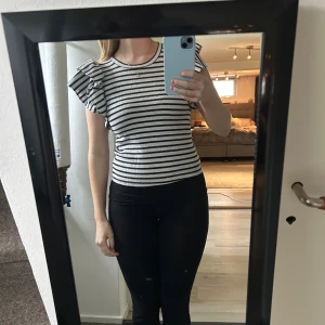 Randig topp med volangärmar - Säljer en svartvit randig topp med volangdetaljer på axlarna. Toppen är ribbad och har en rund halsringning samt korta ärmar med extra volym. Perfekt för dig som gillar en söt men stilren look.