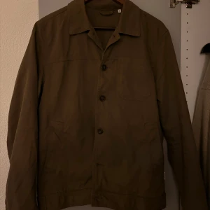 Olivgrön overshirt med knappar - Säljer en stilren olivgrön overshirt med klassisk krage och tre fickor framtill. Jackan har knäppning med knappar och raka ärmar, perfekt för lager-på-lager. Enkel och clean design som passar till det mesta.