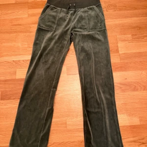Mörkgröna mjukisbyxor Juicy Couture - Mörkgröna mjukisbyxor från Juicy Couture i mjuk sammet med bred resår och snörning i midjan. Byxorna har raka ben, stora fickor fram och bak med knappdetaljer. Perfekta för en chill och avslappnad look.