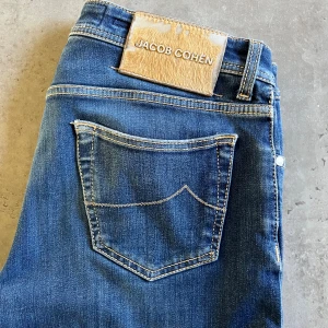 Jacob Cohen jeans - Snygga blå jeans från Jacob Cohen med klassisk femficksdesign och kontrastsömmar. Byxorna har en unik etikett i ljust skinn baktill och knäppning med silverfärgade knappar. Insidan har ett mönstrat tyg vid gylfen för extra detalj. Lite sliten patch och liten defekt mellan benen som syns på bild