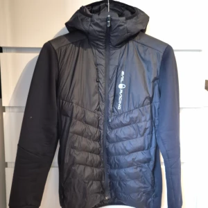 Svart lättviktsjacka från Sail Racing - Svart löttvktsjacka från Sail Racing med huva och dragkedja framtill. Jackan har quiltade partier på framsidan och släta ärmar i mjukt material. Logga tryckt på bröstet. Ficka på bröstet.Perfekt för kyliga dagar och snygg till streetwear. Mindre noppor finns på armar, bra skick. Storlek S