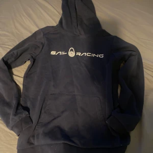 Mörkblå Sail Racing hoodie med tryck - Mörkblå hoodie från Sail Racing med vit logga framtill. Klassisk känguruficka och huva, perfekt för chill dagar. Mjuk insida och ribbade muddar vid ärmslut och nederkant. Snygg och enkel stil som funkar året runt.
