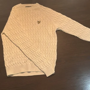 Beige kabelstickad tröja Lyle & Scott - Kabelstickad beige tröja från Lyle & Scott med deras klassiska gula logga på bröstet. Tröjan har rund hals, ribbade muddar och lång ärm. Perfekt för lager-på-lager och snygg till jeans eller chinos. Den är färgen grädd vit