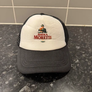 Birra Moretti svartvit truckerkeps - Snygg truckerkeps med svart skärm och mesh samt vit frontpanel med broderad Birra Moretti-logga. Klassisk böjd skärm och justerbar baksida. Perfekt statement-piece för dig som gillar streetwear och italiensk vibe.