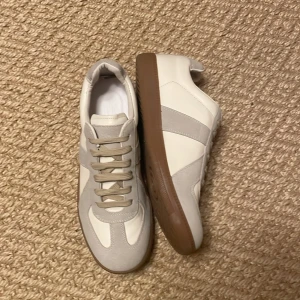 Beige och vita sneakers med mockadetaljer - Snygga sneakers i beige och vitt med detaljer i mocka och skinn. Skorna har en klassisk design med bruna gummisulor och snörning framtill. Perfekta för dig som gillar stilrena och tidlösa sneakers med en modern touch.