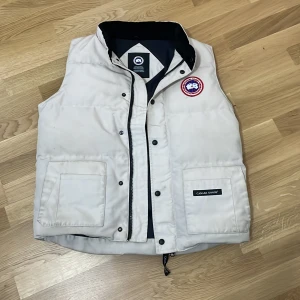 Vit dunväst från Canada Goose - Snygg vit dunväst från Canada Goose med klassisk logga på bröstet och två stora fickor framtill. Västen har dragkedja och knappar, samt svart innerfoder. Perfekt för lager-på-lager och riktigt clean look.