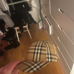 Burberry rutig keps i bomull - Snygg keps från Burberry i klassiskt beige rutigt mönster med svarta, vita och röda detaljer. Justerbar rem med guldfärgat spänne bak. Kepsen är tillverkad i bomull och har Burberry-logga på insidan. Ovanlig modell som är svår och hitta, pris går att diskuteras och säg till ifall fler bilder vill ses av varan😄