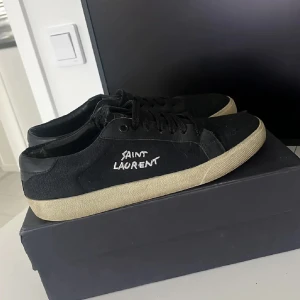 Svarta Saint Laurent sneakers - Svarta sneakers från Saint Laurent med vit broderad logga på sidan. Skorna har svart snörning, rund tå och beige platt gummisula. Perfekta för dig som gillar en clean och stilren look med en touch av lyx. Skorna är storlek 42 och passar även i 43. Mer bilder finns att kolla på. Fraktar samma dag som köp!! Här av er vid frågor eller funderingar. Kan gå ner i pris vid snabb affär!!