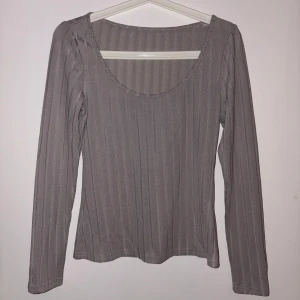 Grå ribbad långärmad topp - En stilren grå långärmad topp med ribbat tyg och rundad urringning. Toppen har en slim passform och är tillverkad i ett mjukt, stretchigt material som sitter snyggt på kroppen. Perfekt att matcha med jeans eller kjol för en enkel men trendig look.